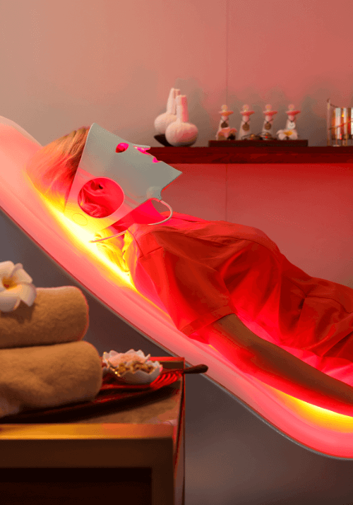 Woman wearing a light therapy face mask while relaxing in the Energy Lounger with towels in a spa or wellness room.