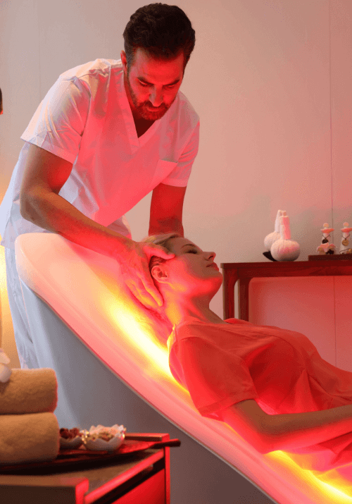 Spa therapist providing a relaxing head massage to a person lying on an Energy Lounger red-light therapy bed.