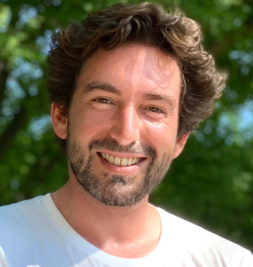 A smiling man with curly hair and a beard standing outdoors with green trees in the background.