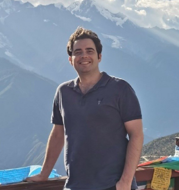 A man standing outdoors in front of mountains, smiling with a relaxed posture.