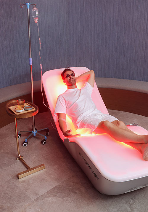 Man relaxing on an Energylounger chair receiving red light therapy beside an IV stand in a wellness room.