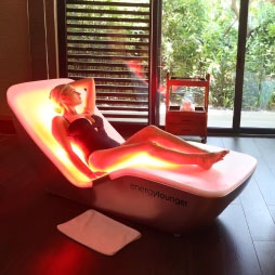 Woman relaxing in a red-light therapy Energy Lounger placed indoors with a view of an outdoor garden.